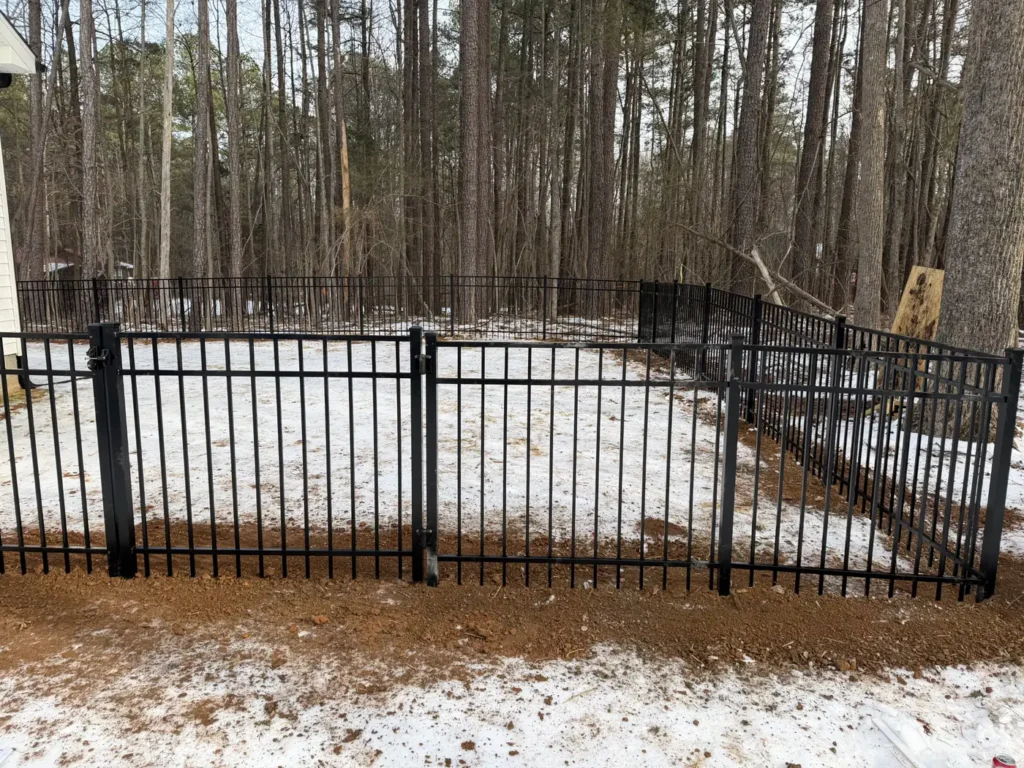 Black aluminum fence and gate in a snowy yard lined with pine trees