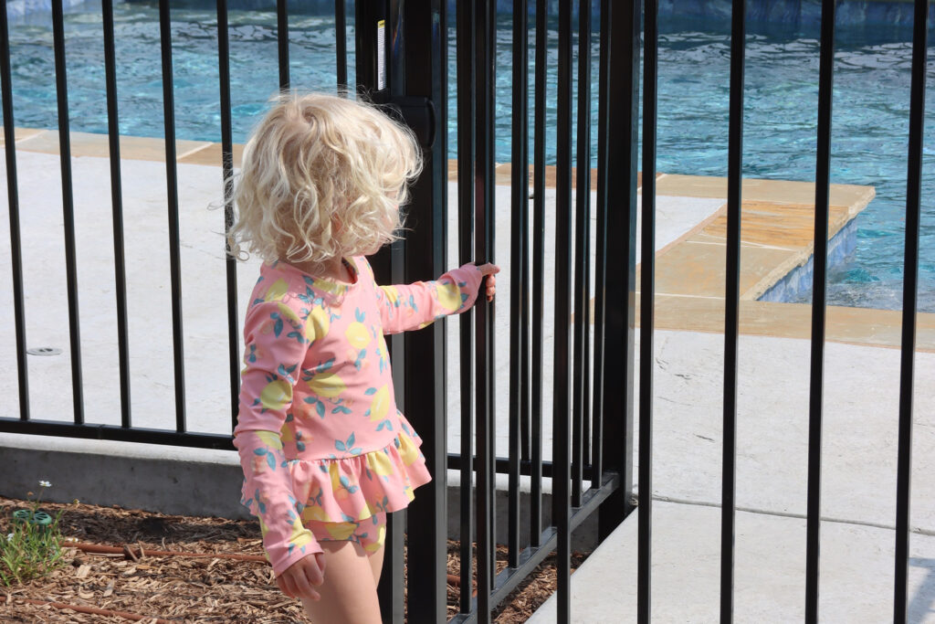 Young girl standing at a black metal pool fence beside a swimming pool