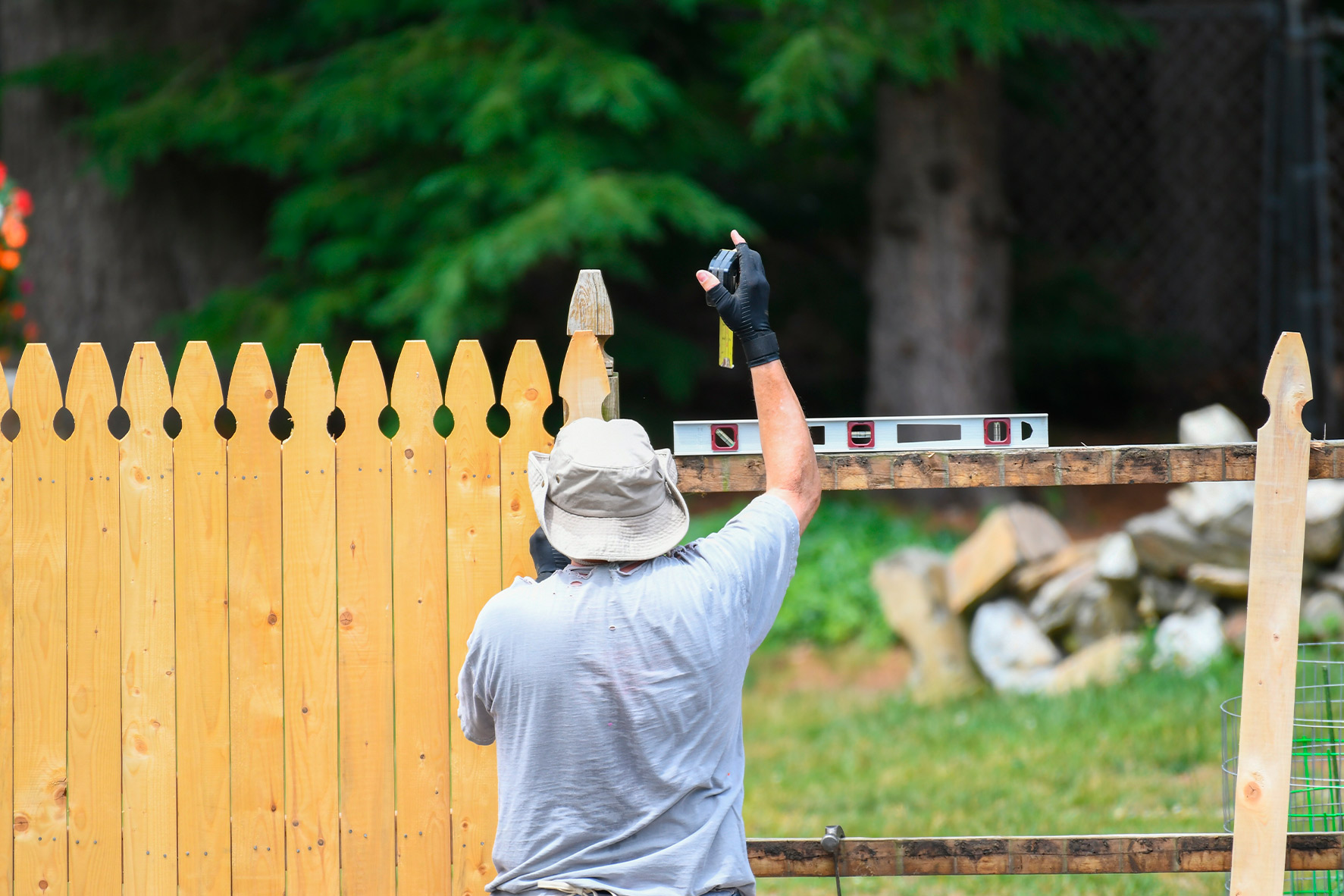 A man is engaged in building a fence, carefully measuring and securing the wooden panels in place.