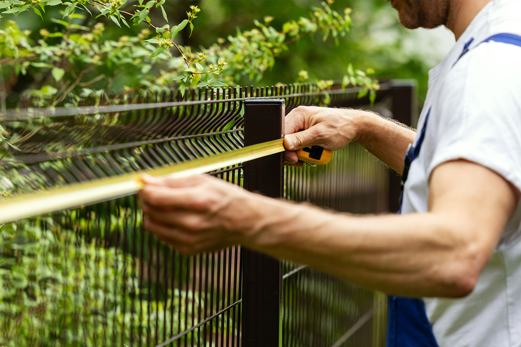 A man in a white shirt and blue overalls measures a metal fence with a yellow tape measure, surrounded by green foliage, conveying focus and precision.