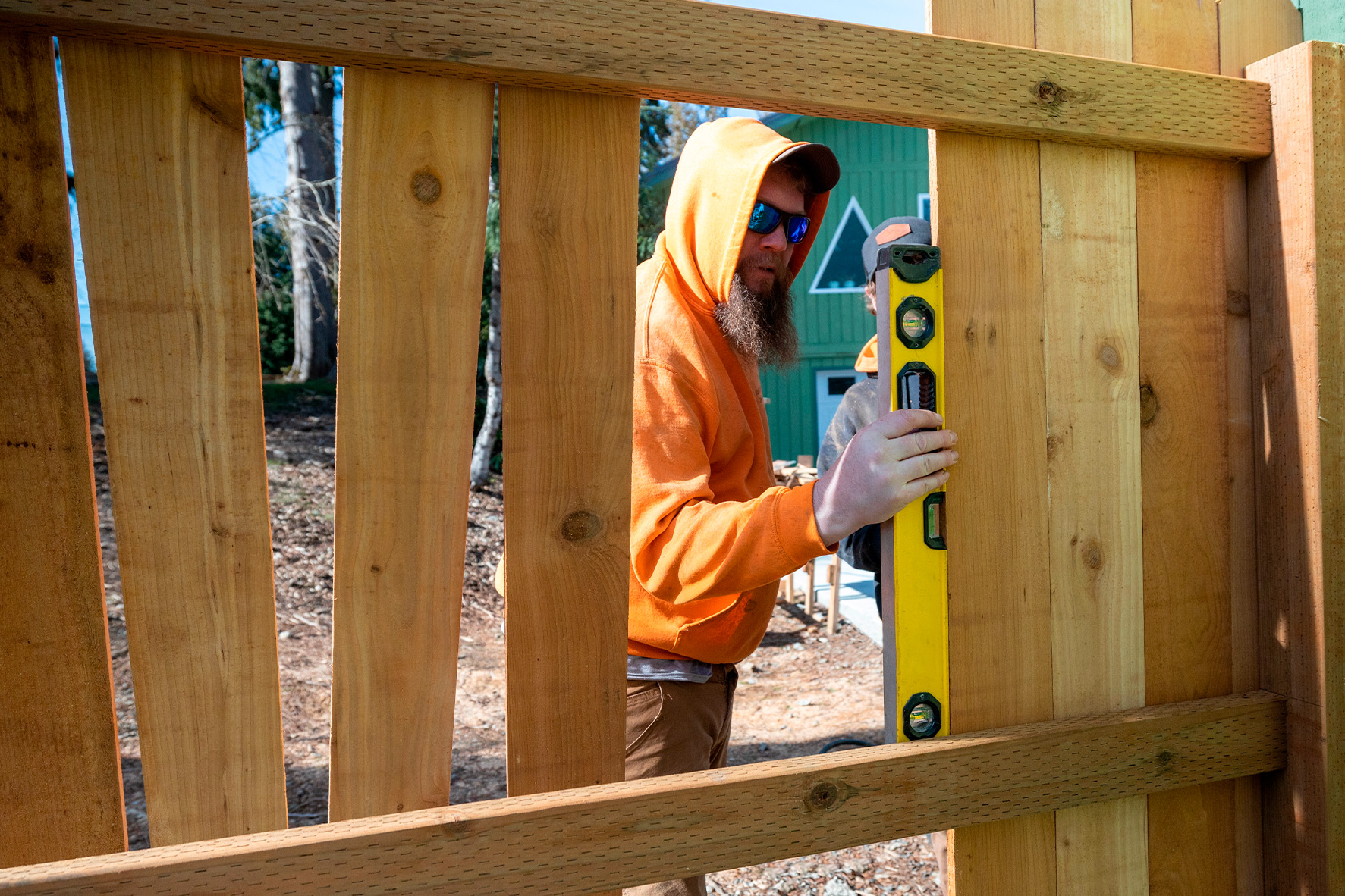 A man in an orange hoodie poses beside a wooden fence, relevant to the wood fence repair cost guide.
