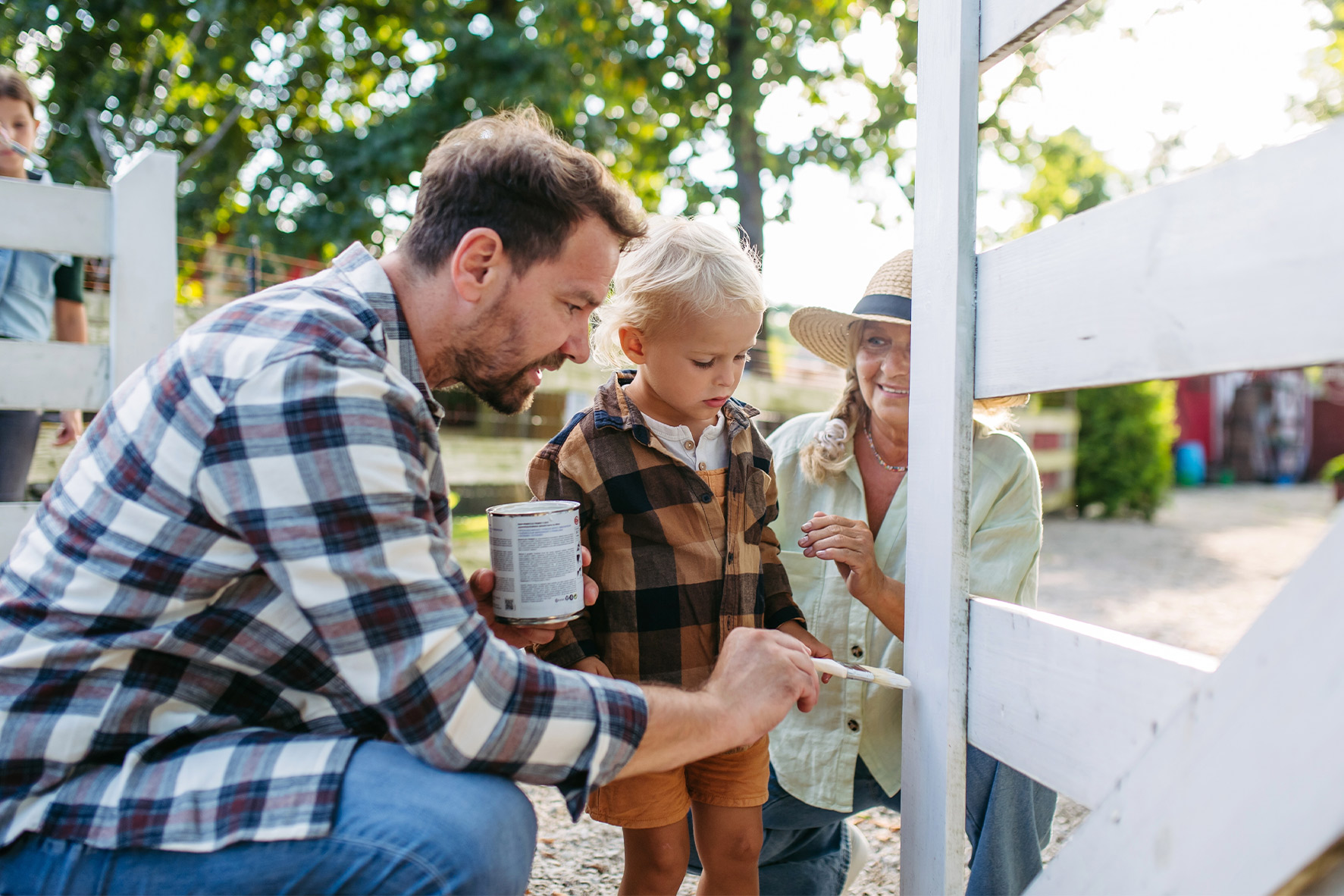 A man and woman are collaboratively painting a fence while a child assists them, surrounded by greenery.