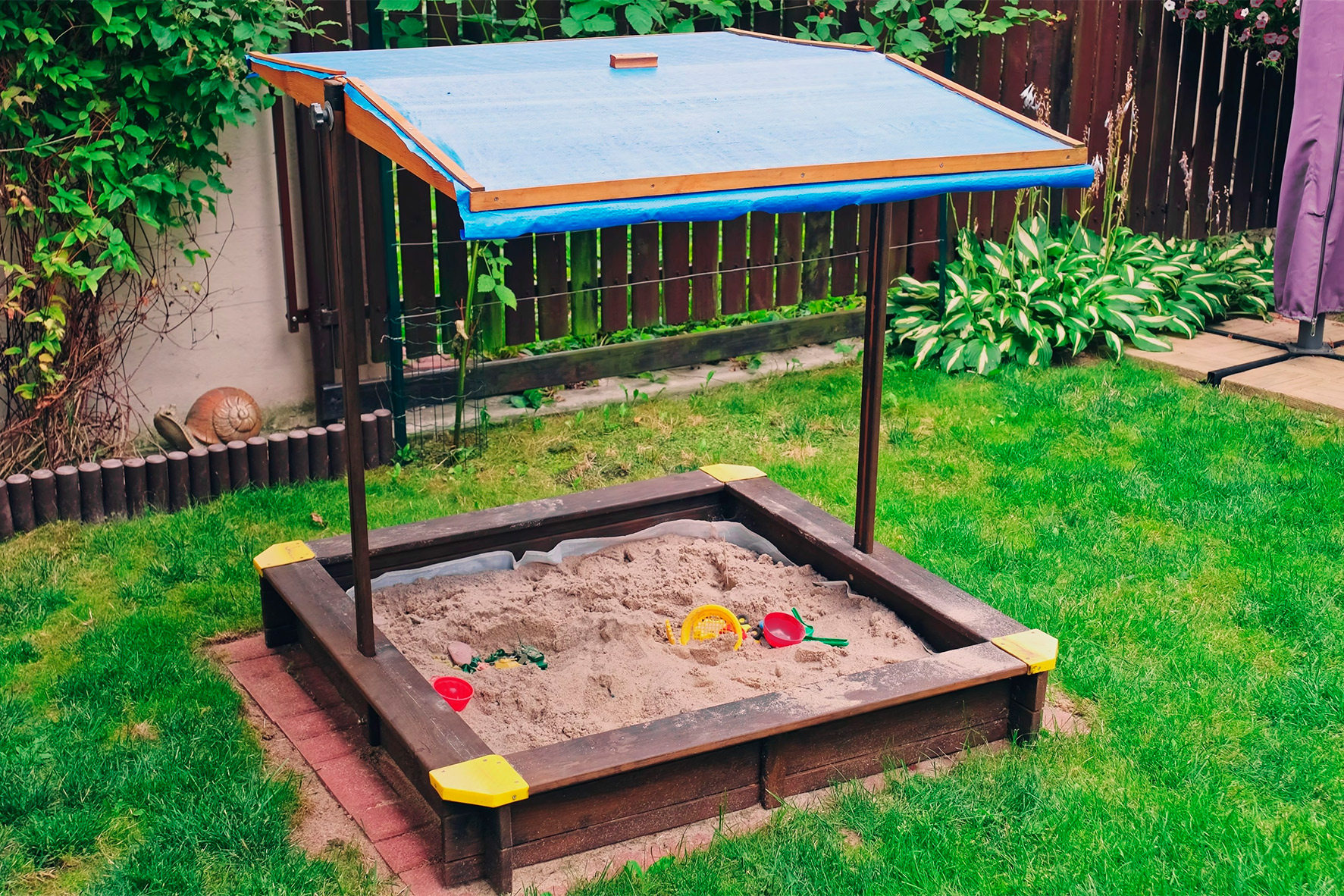  A sandbox covered by a blue canopy, providing shade for children playing inside. Fence around the backyard