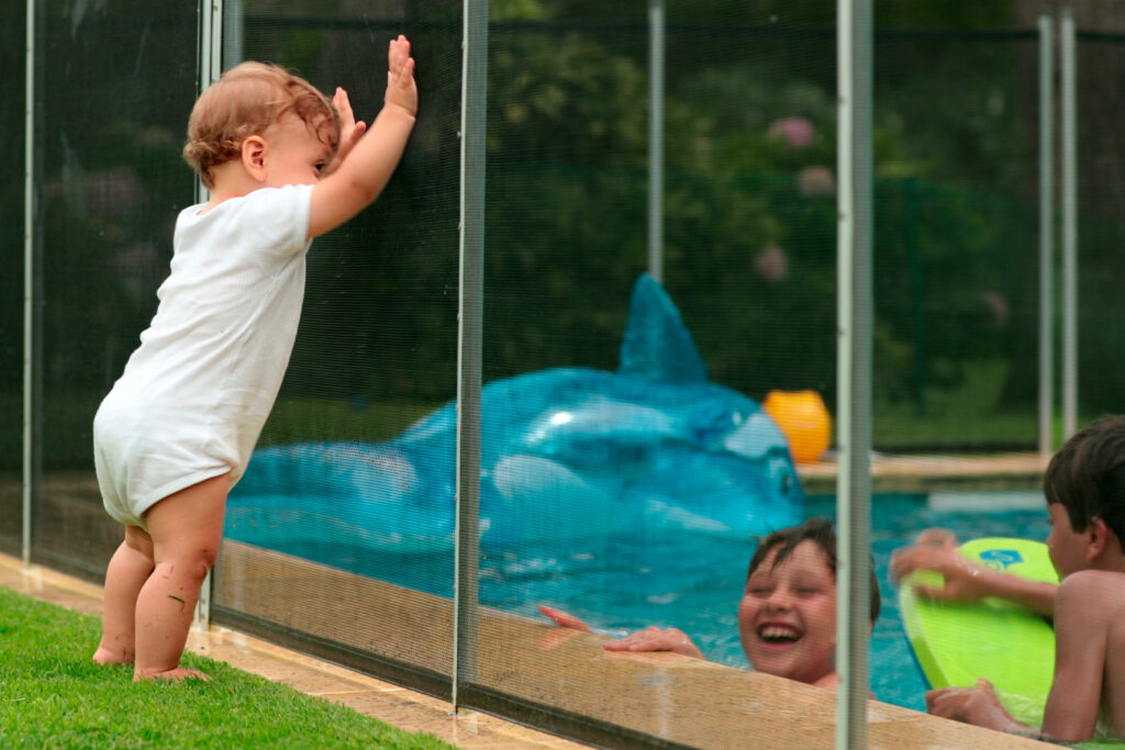 Toddler standing against a mesh pool fence while children swim on the other side
