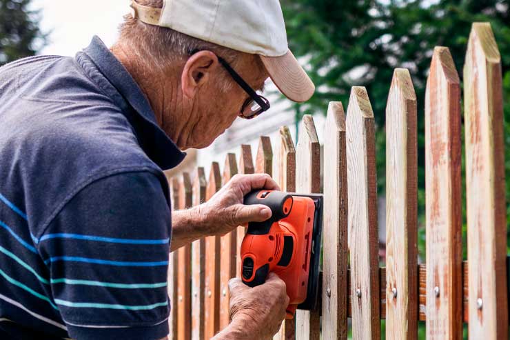 Sanding and preparing wooden fence for painting. Senior man restoring picket fence in garden