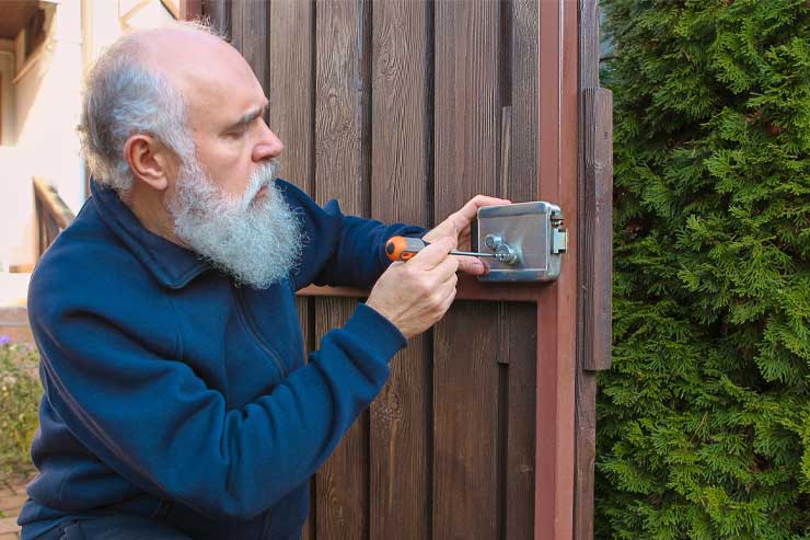 Senior man adjusting fence lock with concentrated precision during home repair. Concept of DIY tasks, outdoor maintenance, security hardware visuals, and practical homeowner routines.