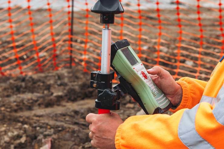 Surveyor holding a GPS survey pole and data collector beside orange safety fencing