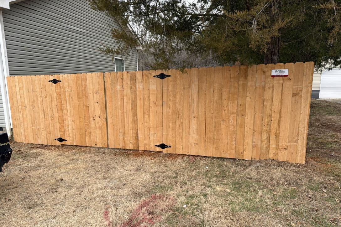 A tall wood fence stands in a yard, bordered by a house and tree, providing privacy and a natural rustic look to the setting.