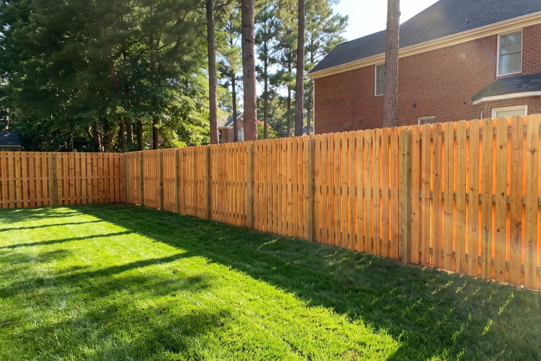 Wooden fence encloses a lush green garden in NC, with sunlight casting shadows. Brick house and tall trees in the background.