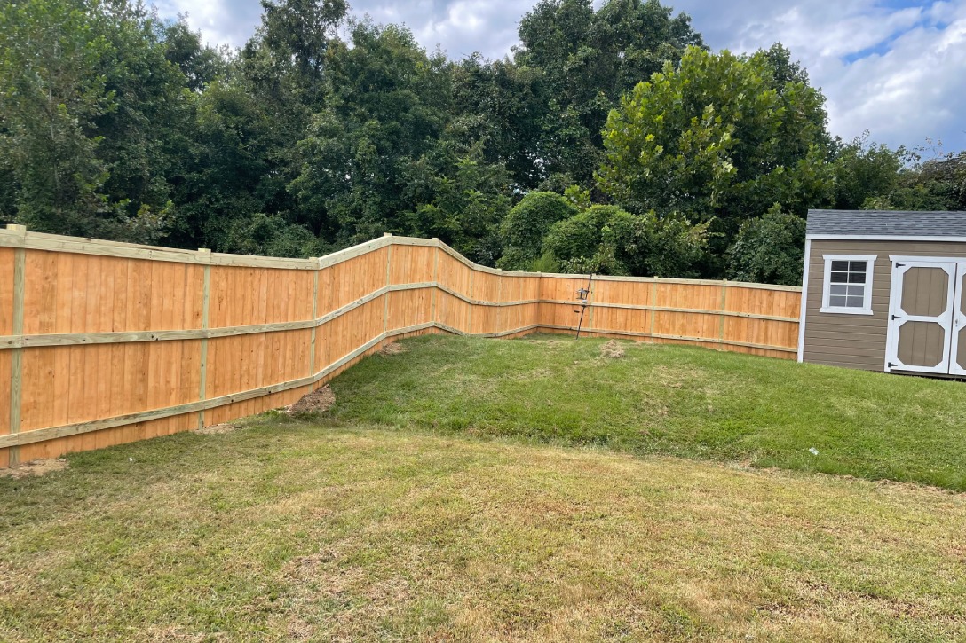 A wooden fence encloses a backyard featuring a small shed, surrounded by grass and plants.